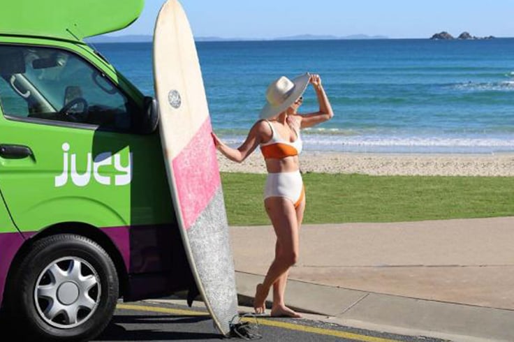 Girl at the beach with a surfboard and a JUCY campervan