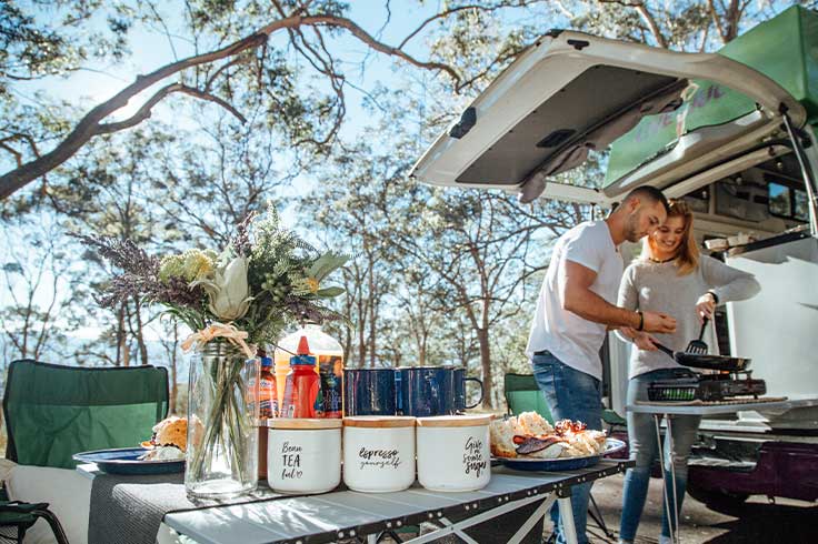A couple cooking out of their JUCY Condo Campervan kitchen with a dinner table set up